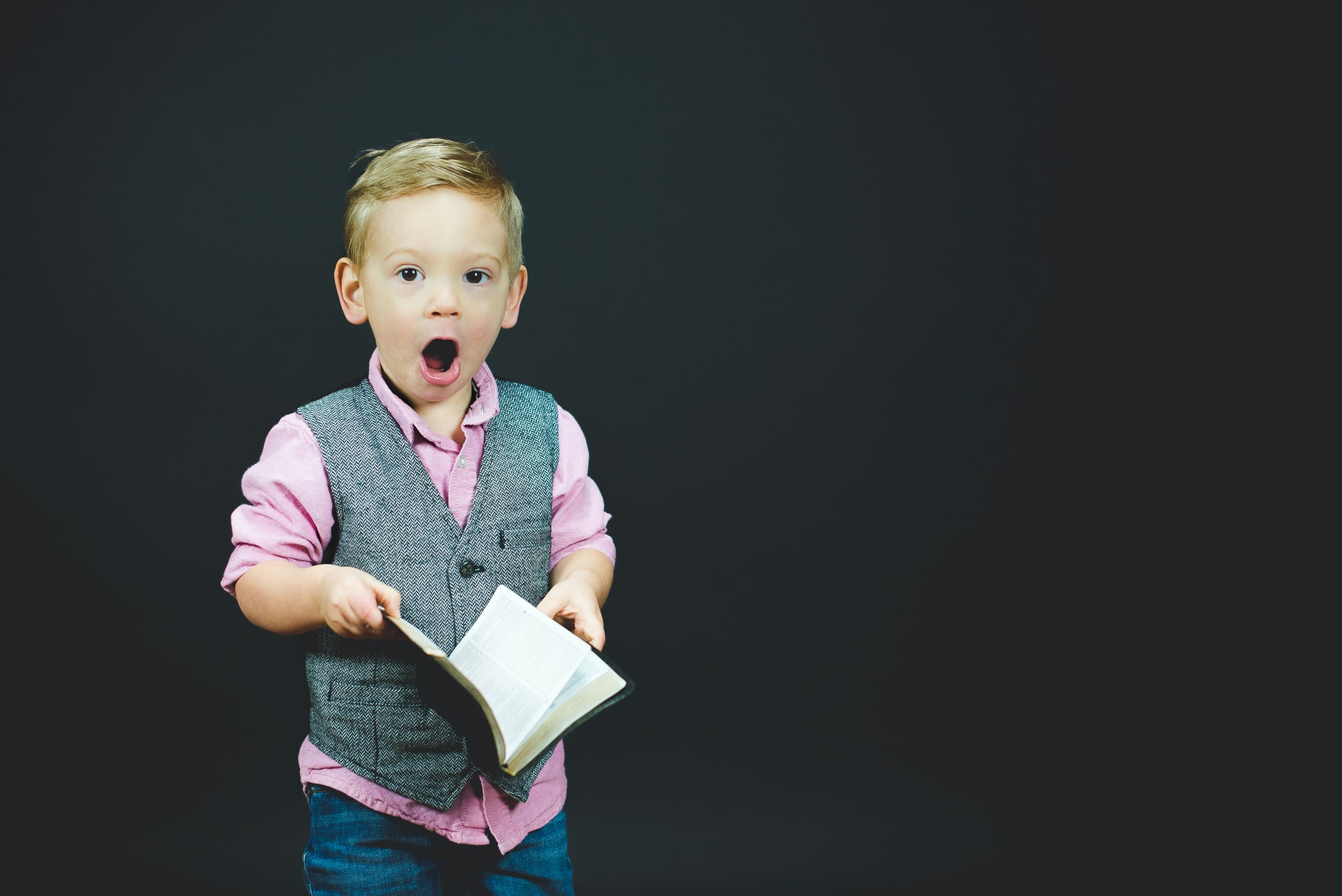 Boy in pink shirt with gray vest reacts with shock to open book in his hands