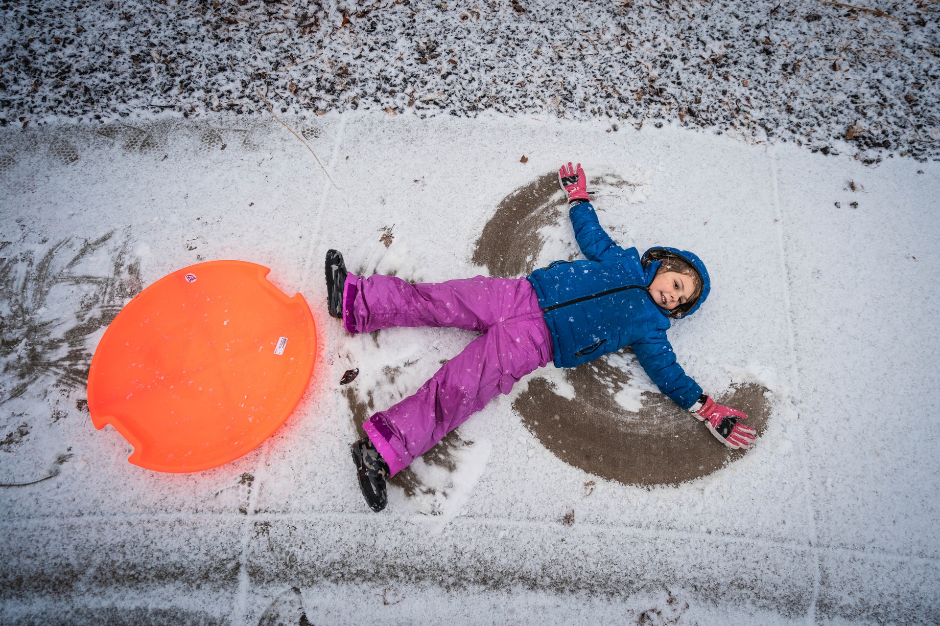 Girl making snow angel on sidewalk next to orange circular sled
