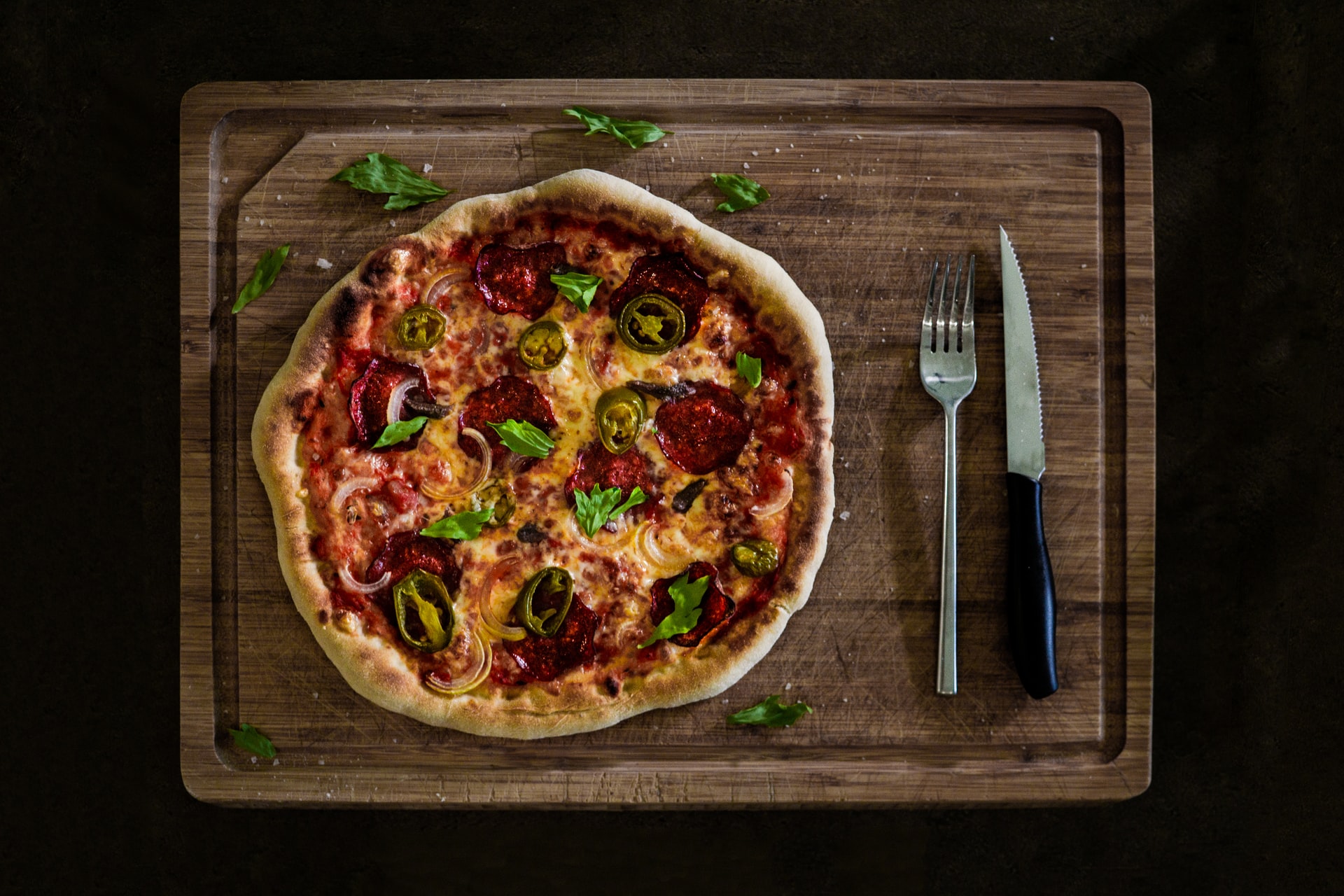 Overhead on gourmet pizza with jalapenos, pepperoni, and onions served on wooden cutting board with knife and fork next to it